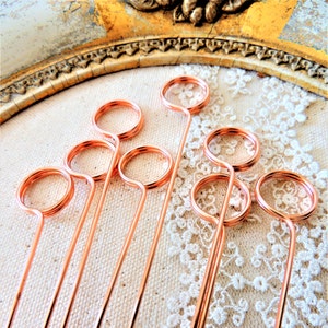 May include: A set of eight rose gold wire place card holders. Each holder has a small loop at the top and a long, thin stem.