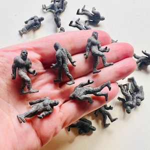 May include: A collection of small, dark gray plastic figurines depicting a hairy, bipedal creature in various poses. Each miniature figure features textured details representing fur, with some standing upright and others lying down.