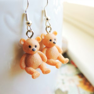 May include: A pair of dangle earrings featuring small, brown teddy bear charms. The bears have black eyes and noses.