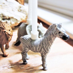May include: A set of animal candle holders featuring a zebra, a lion, and a rhinoceros. The candle holders are made of a white ceramic material and are designed to hold small candles.