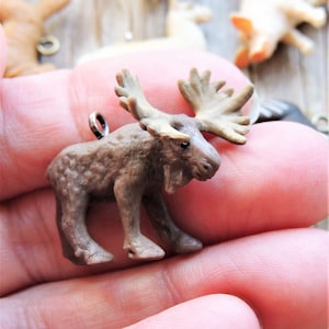 May include: A small brown moose figurine with large antlers. The figurine is made of plastic and has a brown and white color scheme. It is a charm or pendant with a metal loop for hanging.