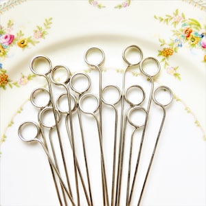 May include: A set of silver metal skewers with round loops at the top. The skewers are arranged in a fan shape on a white plate with a floral pattern.