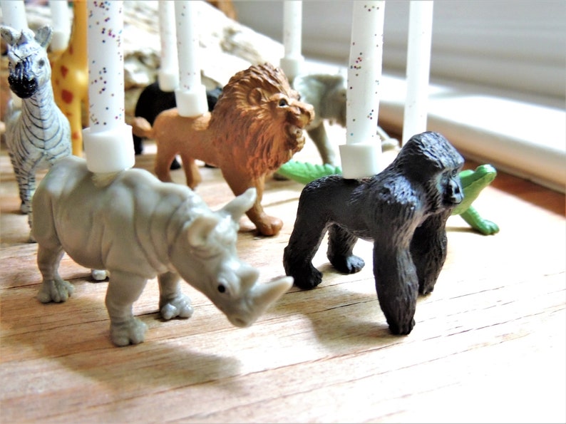 May include: A set of animal figurines with white candles, perfect for a birthday cake. The figurines include a zebra, giraffe, lion, rhinoceros, gorilla, and a green alligator.