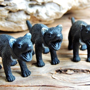 May include: Three black toy Tasmanian devils with open mouths, standing on a wooden surface. The figures have detailed features, including visible teeth and claws. The background features a piece of driftwood.