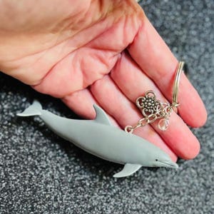 May include: A gray dolphin keychain with a silver-colored keyring and charms. The dolphin has a detailed design and is attached to a chain with a flower charm and a clear crystal bead. The keychain is held in a hand.