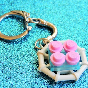 May include: A white plastic keychain with a silver ring. The keychain has four pink Lego bricks in a square shape.