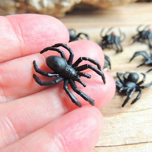 May include: A collection of small, black plastic spider figurines. Each spider features a rounded abdomen, a distinct cephalothorax, and eight articulated legs. The surface of the spiders is smooth with a matte black finish, showing molded details on the body and legs.