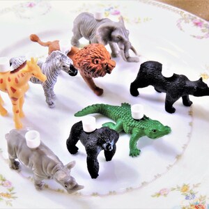 May include: A set of nine plastic animal figurines, each with a white plastic ring on top. The animals include a giraffe, zebra, lion, elephant, bear, alligator, and rhinoceros. The figurines are arranged on a white plate with a floral pattern.