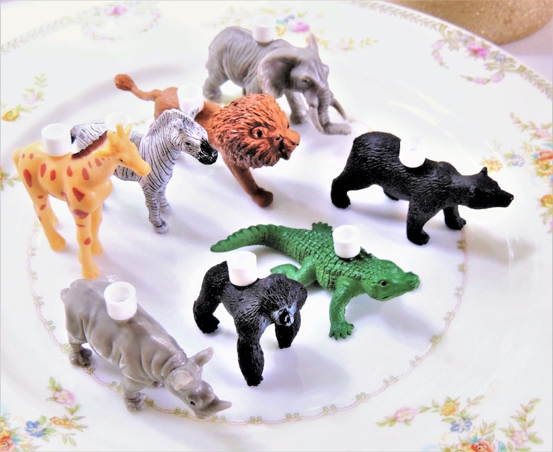 May include: A set of nine plastic animal figurines, each with a white plastic ring on top. The animals include a giraffe, zebra, lion, elephant, bear, alligator, and rhinoceros. The figurines are arranged on a white plate with a floral pattern.