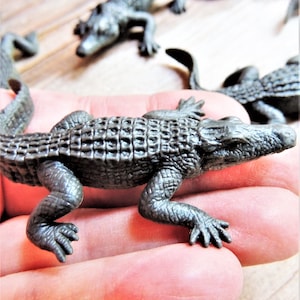 Peut inclure: Plusieurs petites figurines d'alligators en plastique gris. L'une est mise au point, montrant des écailles et des pattes détaillées. D'autres alligators sont en arrière-plan, légèrement flous. Les alligators sont sur une surface claire.
