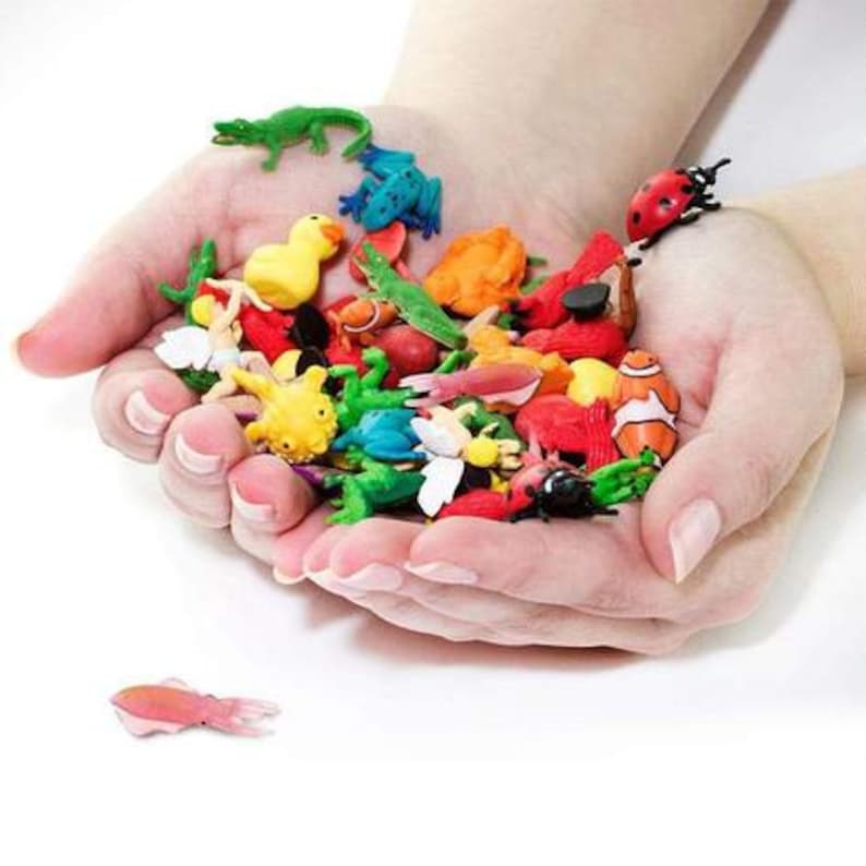 May include: A collection of colorful plastic animal figurines, including a frog, a ladybug, a fish, a turtle, a butterfly, and a lizard. The figurines are arranged in a person's hands.