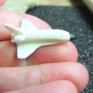 Könnte beinhalten: Ein kleines, weißes Spielzeug-Space Shuttle mit schwarzen Details an Nase und Flügeln. Das Miniatur-Raumschiff wird zwischen Fingern gehalten, was seine kompakte Größe und sein Design hervorhebt. Das Spielzeug ist eine Miniatur-Nachbildung.