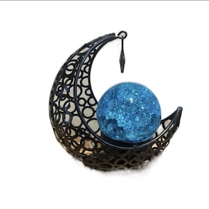 May include: A black metal crescent moon sculpture featuring an intricate, openwork circular pattern. A round, textured blue glass sphere with a crackled finish is nestled within the curve of the moon, suspended by a small metal hook at the top.