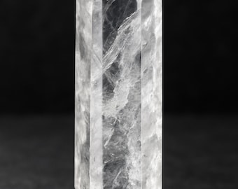 Könnte beinhalten: Ein klarer Quarz-Kristallpunkt mit einer facettierten, länglichen Form. Der Kristall ist transparent mit inneren Adern und verjüngt sich an beiden Enden zu einer scharfen Spitze. Der Kristall wird vor einem dunklen Hintergrund gezeigt.