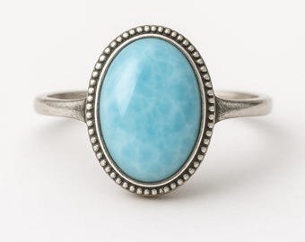 May include: A silver ring with an oval, pale blue gemstone. The gemstone is framed by a beaded edge. The ring has a simple, elegant design, suitable for daily wear. The ring is set against a white background.