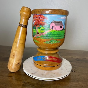 May include: Wooden mortar and pestle with a painted scene of a house and trees. The mortar has a blue, red, and white ribbon around the base with the text "Rep. Dominicana".
