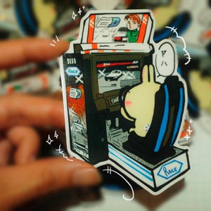 May include: A sticker featuring a cartoon arcade game with a bunny character playing a racing game. The arcade cabinet is black with the Sega Race logo. The bunny is in a black and blue seat, with a thought bubble above its head.