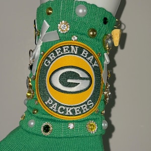 May include: A bright green knit sock featuring a large embroidered Green Bay Packers logo patch in yellow, green, and white. The sock is embellished with various decorative elements including white satin bows, clear rhinestones, pearls, and a small yellow cheese wedge charm.