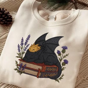 May include: Cream-colored sweatshirt featuring an embroidered design of a sleeping black dragon with gold accents, resting on two books. Lavender and thistle flowers surround the dragon. A cozy, fantasy-themed garment.