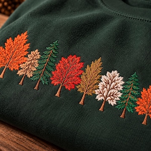 Op de afbeelding: Een donkergroene sweater met ronde hals en een geborduurde rij van diverse bomen op de borst. Het ontwerp bevat esdoorns, dennen en loofbomen in tinten oranje, rood, bruin, crème en groen, uitgevoerd met gedetailleerd stikwerk.