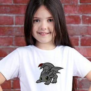 May include: White t-shirt featuring a cartoon dragon in shades of gray with green eyes and red accents. The dragon is in a crawling position. The shirt is made of a soft material and is suitable for children.