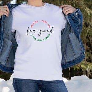 Puede incluir: Sudadera blanca de cuello redondo con la frase "for good" en cursiva, rodeada por las frases "Because I knew you" e "I have been changed". El texto es en negro, rojo y verde.