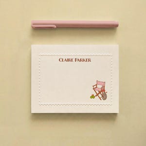 May include: A cream-coloured rectangular note card with a scalloped border, featuring the name CLAIRE PARKER printed in brown and a small illustration of a pink and white striped director's chair, accompanied by a matching dusty pink pen.