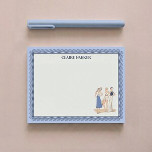 Può includere: Un blocco note azzurro con un bordo decorativo blu scuro e il nome Claire Parker stampato in alto. L'angolo in basso a destra presenta un'illustrazione di tre figure in abiti vintage. Una penna azzurra abbinata è posizionata sopra.