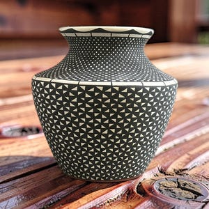 Puede incluir: Un pequeño jarrón de cerámica con acabado negro mate y complejos patrones geométricos blancos. El diseño incluye una cuadrícula repetitiva de pequeños triángulos en el cuerpo, una banda punteada en el cuello y un borde blanco festoneado.