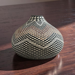 May include: A rounded ceramic vase featuring a complex geometric pattern of black, white, and tan squares and triangles. The vessel has a narrow opening at the top and a wide, bulbous body that tapers toward the base, covered in a repeating grid design.