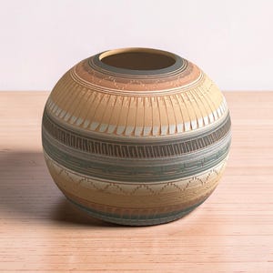 May include: A spherical ceramic vase featuring a matte finish with horizontal bands of earth tones including tan, beige, grey, and muted blue. The surface is decorated with intricate carved geometric patterns, including zigzags and lines.