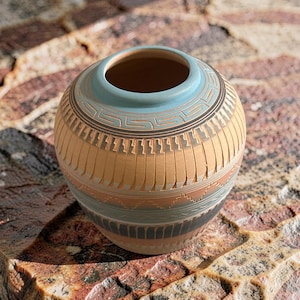 Puede incluir: Un jarrón redondo de cerámica artesanal, con cuerpo marrón claro y borde azul claro. El jarrón presenta intrincados patrones geométricos en tonos marrón, gris y azul. La cerámica es probablemente hecha a mano y adecuada para la decoración del hogar.