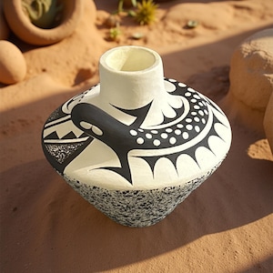 May include: A handcrafted ceramic vase with a unique design. The vase features a cream-colored body with black geometric patterns and a textured, speckled base. The design includes a stylized whale or fish motif. The vase is displayed on a sandy surface.
