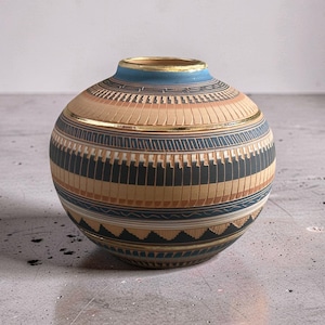 May include: A spherical ceramic vase featuring a matte finish with horizontal bands of tan, dark grey, and blue. The surface is decorated with intricate geometric patterns, including zigzags and lines, accented by a thin gold rim at the opening.