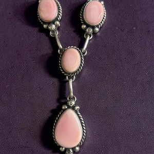 Pode incluir: Um colar prateado com uma série de pedras ovais e em forma de gota rosa. As pedras são colocadas numa moldura prateada decorativa com um design de corda torcida. O colar tem um design em forma de Y.