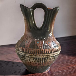 May include: A ceramic vase with a double-spout handle design, featuring a rounded body and a tapered neck. The surface displays a gradient of dark brown and earthy orange tones with etched geometric patterns and small, inlaid turquoise-colored dots.