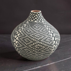 May include: A handcrafted ceramic vase with a rounded body and a narrow neck. The vase features a black and white geometric pattern of triangles and rectangles. The interior of the neck is brown. The vase is displayed on a dark surface.
