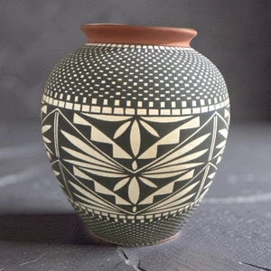 May include: A rounded ceramic vase featuring a matte black finish with intricate white geometric patterns. The design includes a stippled dot texture at the top and bottom, with a central band of repeating angular floral and zigzag motifs.