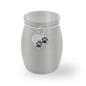 May include: A silver metal urn with a small heart-shaped charm with paw prints. The urn is for pet ashes.