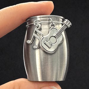 May include: A small, cylindrical, brushed silver metal container featuring a thin wire rim around the top. Two silver-toned charms are attached to the wire: one shaped like a musical note and the other shaped like an acoustic guitar.