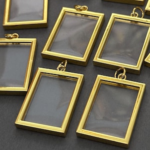 May include: Several small, rectangular gold-colored pendant frames with clear inserts. Each frame has a small loop at the top for attaching to a chain or other jewelry component. The frames are arranged on a dark gray surface.