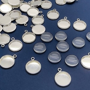 May include: A collection of silver-toned metal pendant trays with round, flat bottoms and a loop at the top. There are also several clear glass cabochons with a slightly textured surface.