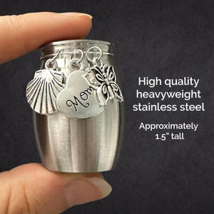 May include: A small, silver-colored stainless steel keepsake urn, approximately 1.5 inches tall. It features three hanging charms: a seashell, a heart engraved with the word Mom, and a butterfly. The text reads High quality heavyweight stainless steel.