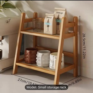 Op de afbeelding: Een kleine, houten opbergrek met twee niveaus, 27 cm breed en 28,5 cm hoog, met een rasterpatroon op de planken. Het bevat koffiezakken, mokken en een glas. De tekst op de zakken luidt "SILVER CUP" en "WONDERLAND".