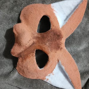 May include: A handmade fox mask with a brown and white color scheme. The mask features eye holes and pointed ears. The mask is made of felt and is designed to be worn on the face.