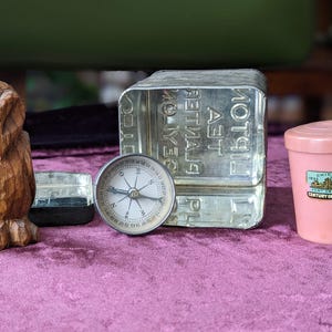 May include: A wooden owl carving, a compass, a metal container, a small pink container with a lid, and a small metal box are arranged on a purple surface. The pink container has a label that says "Chicago 1933 Century of Progress."
