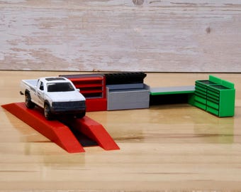 May include: A white pickup truck on a red ramp with a set of toy garage workbenches in the background. The workbenches are red, gray, black, and green.