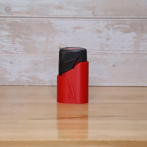 Op de afbeelding: Een rode Milwaukee batterijhouder met een zwarte batterij erin.