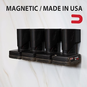 May include: A black magnetic battery holder for power tools, holding four batteries. The text "MAGNETIC / MADE IN USA" is printed at the top. The holder is mounted on a white and beige marble-patterned wall.