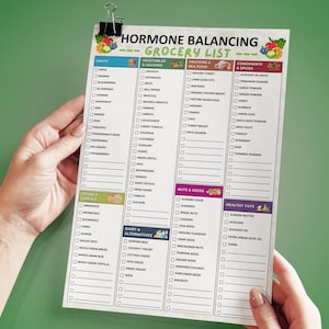 May include: A white grocery list titled "Hormone Balancing Grocery List" held by two hands. The list is divided into sections for fruits, vegetables, proteins, condiments, nuts, and healthy fats. A black clip holds the list.